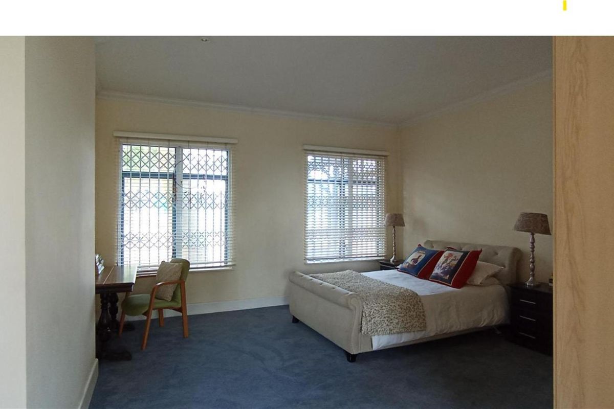 A spacious bedroom featuring neutral walls, carpeted flooring, and two windows fitted with security bars and blinds.