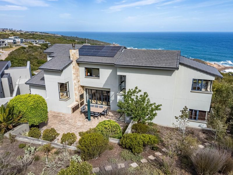 Aerial view of a modern coastal residence featuring a tiled roof with solar panels, stone chimney, and a paved patio overlooking the ocean.