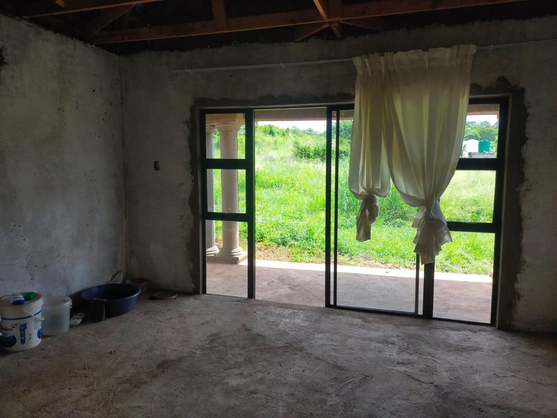An unfinished living area featuring exposed roof trusses, raw masonry walls, and large sliding glass doors opening onto a patio and garden.
