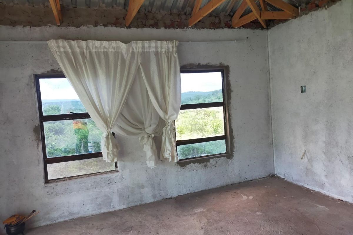 An unfinished bedroom space featuring exposed timber roof trusses, cement screed flooring, and aluminium window frames with views of the surrounding landscape.