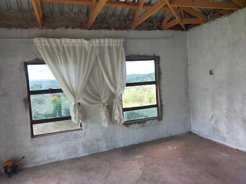 An unfinished bedroom space featuring exposed timber roof trusses, cement screed flooring, and aluminium window frames with views of the surrounding landscape.