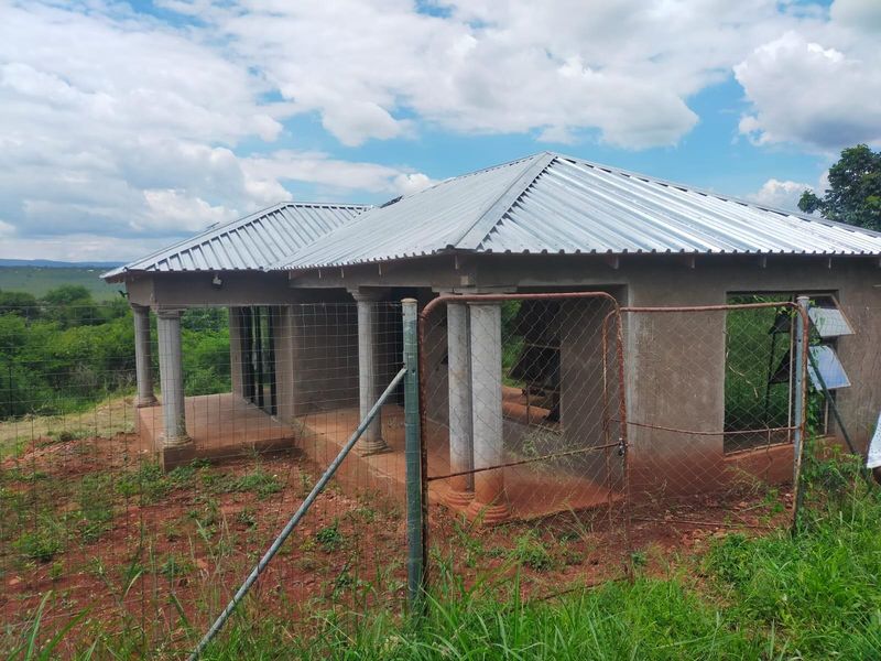 An unfinished single-story house featuring a corrugated metal roof, concrete columns, and a covered veranda, set on a rural plot with wire fencing.