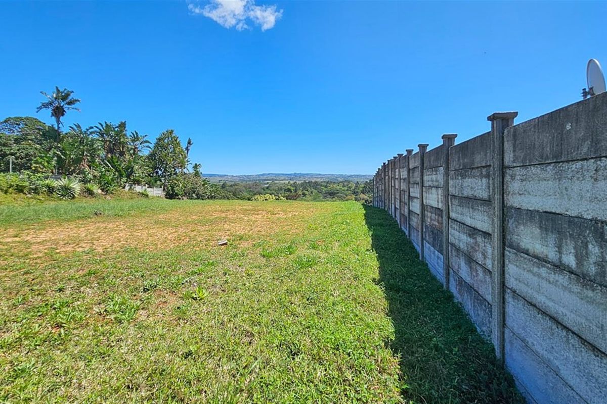 A vacant residential plot featuring a grassed surface and a long concrete boundary wall, offering expansive views of the surrounding landscape.