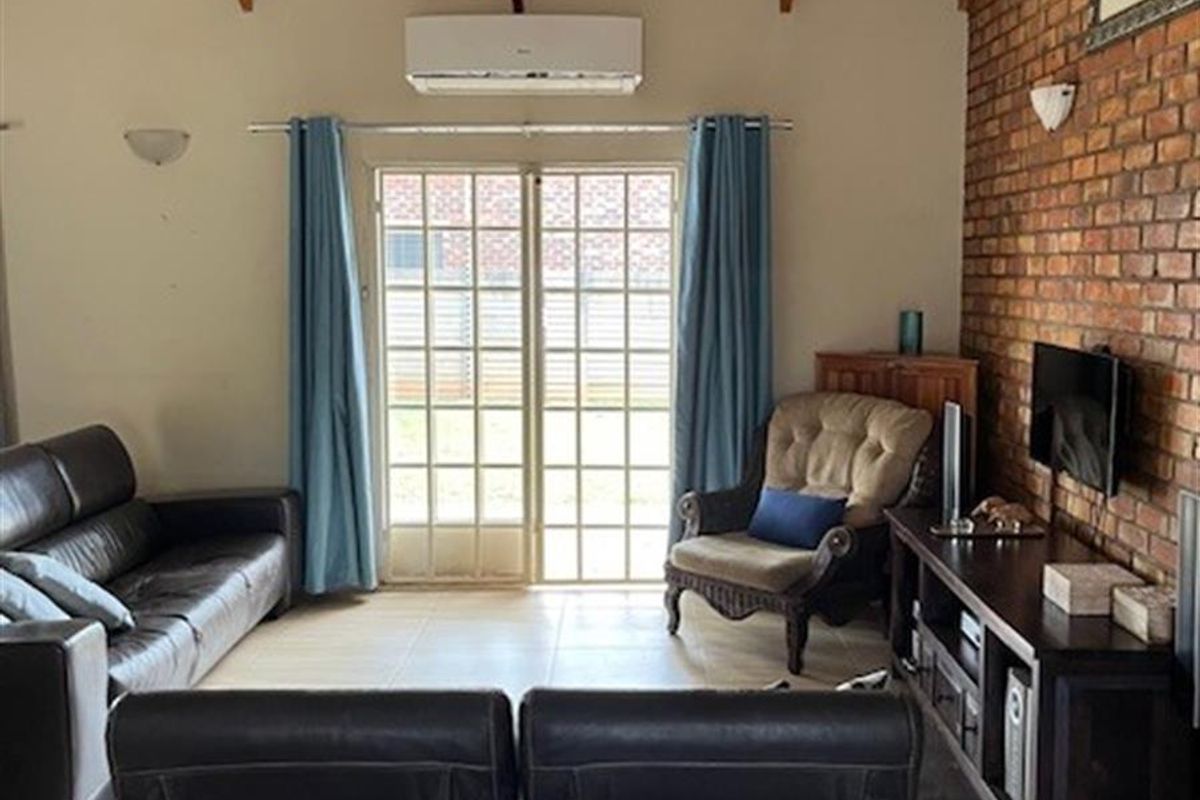 A living room featuring a vaulted ceiling with exposed timber beams, a feature face-brick wall, tiled flooring, and a wall-mounted air conditioning unit.
