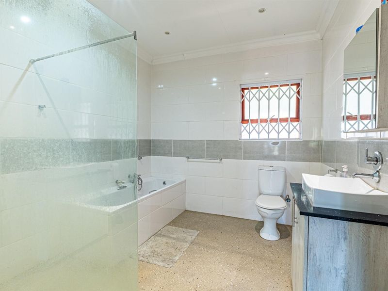 A functional bathroom featuring a built-in bathtub, glass shower screen, vessel sink vanity, and a window with security bars.
