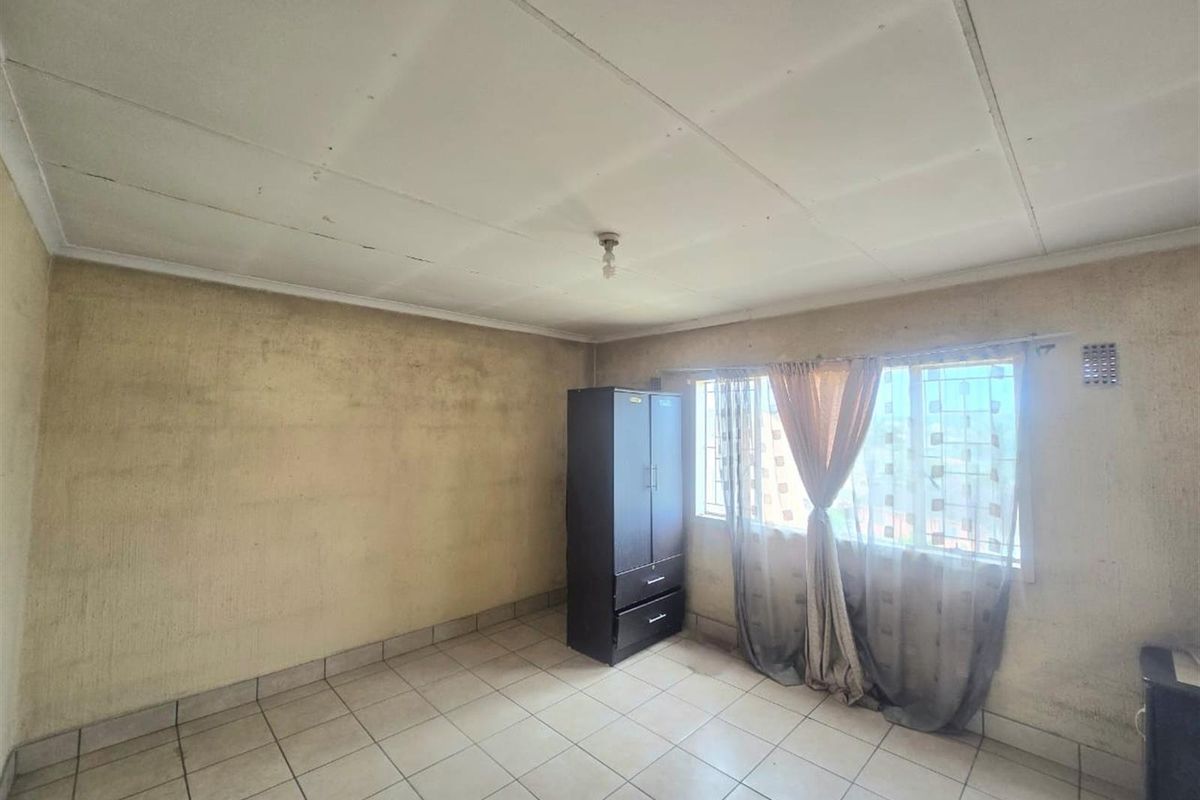 A bedroom featuring tiled flooring, a window with security bars, and a ceiling light fixture. The room requires significant cosmetic updates and wall repairs.