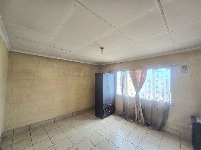 A bedroom featuring tiled flooring, a window with security bars, and a ceiling light fixture. The room requires significant cosmetic updates and wall repairs.