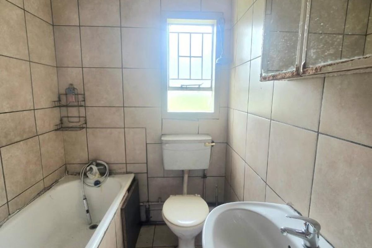 A dated bathroom featuring a standard bathtub, pedestal basin, and toilet, with floor-to-ceiling wall tiling and a window for natural light.