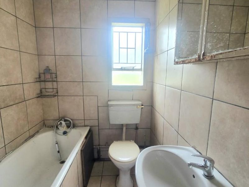 A dated bathroom featuring a standard bathtub, pedestal basin, and toilet, with floor-to-ceiling wall tiling and a window for natural light.