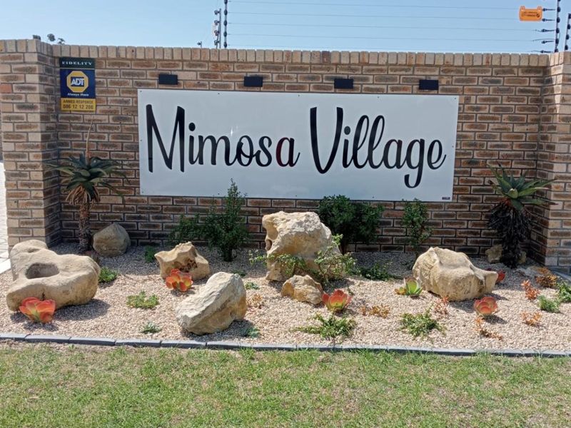 Entrance signage for Mimosa Village featuring a brick boundary wall, security fencing, and landscaped rock garden.