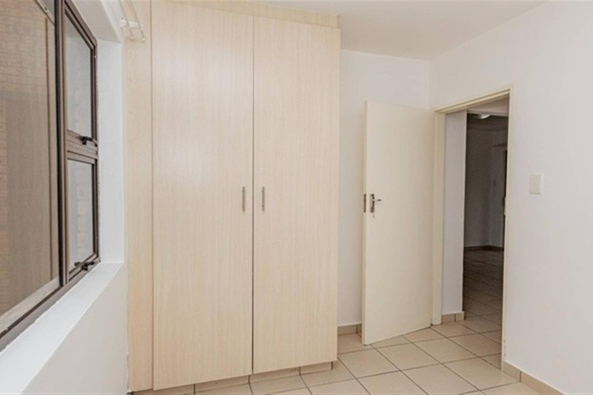A bedroom featuring built-in light wood-finish cupboards, tiled flooring, and a window overlooking an exterior brick wall.
