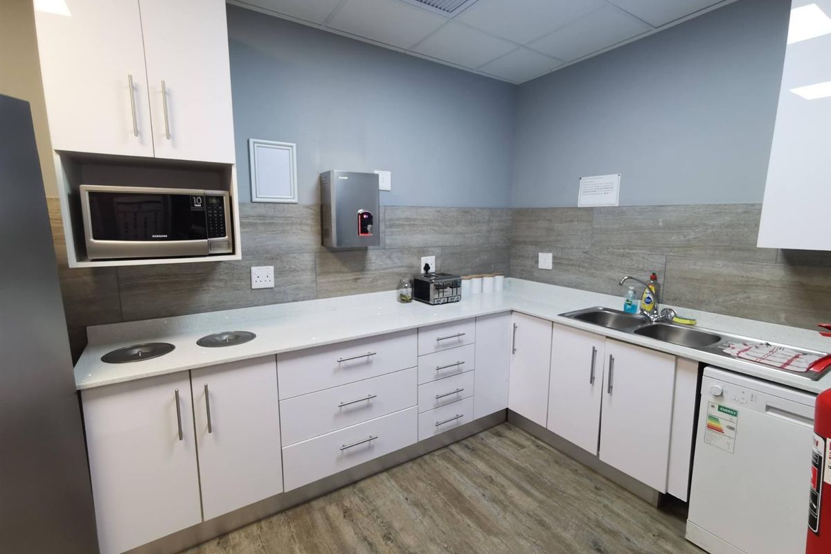 A clean, modern commercial kitchenette featuring white cabinetry, stone-look countertops, a double stainless steel sink, and integrated microwave.