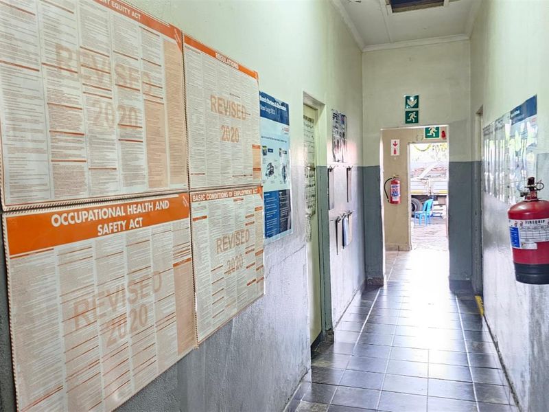 A functional office hallway featuring tiled flooring, wall-mounted safety signage, and a fire extinguisher, leading towards an exterior exit.