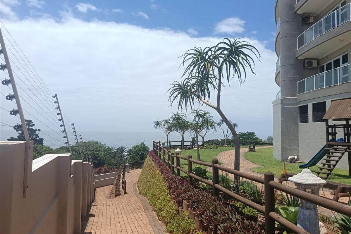 A well-maintained rear garden area featuring a paved walkway, timber fencing, and landscaped grounds with ocean views adjacent to the penthouse building.