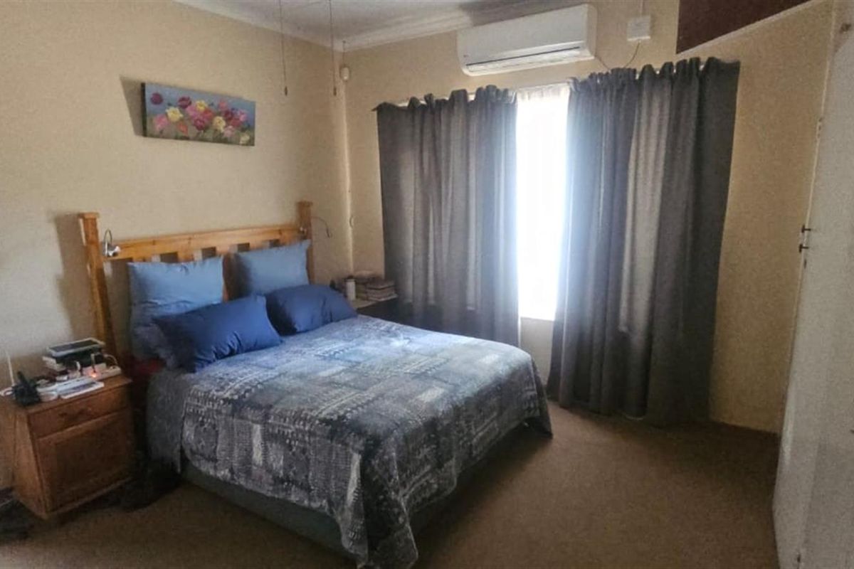 A standard bedroom featuring carpeted flooring, a wall-mounted air conditioning unit, and a window with curtain treatments.