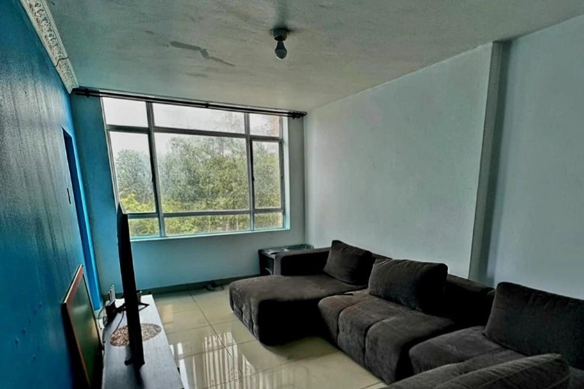 A living room featuring large windows, tiled flooring, and decorative ceiling cornicing, currently showing signs of ceiling water damage and uneven wall finishes.
