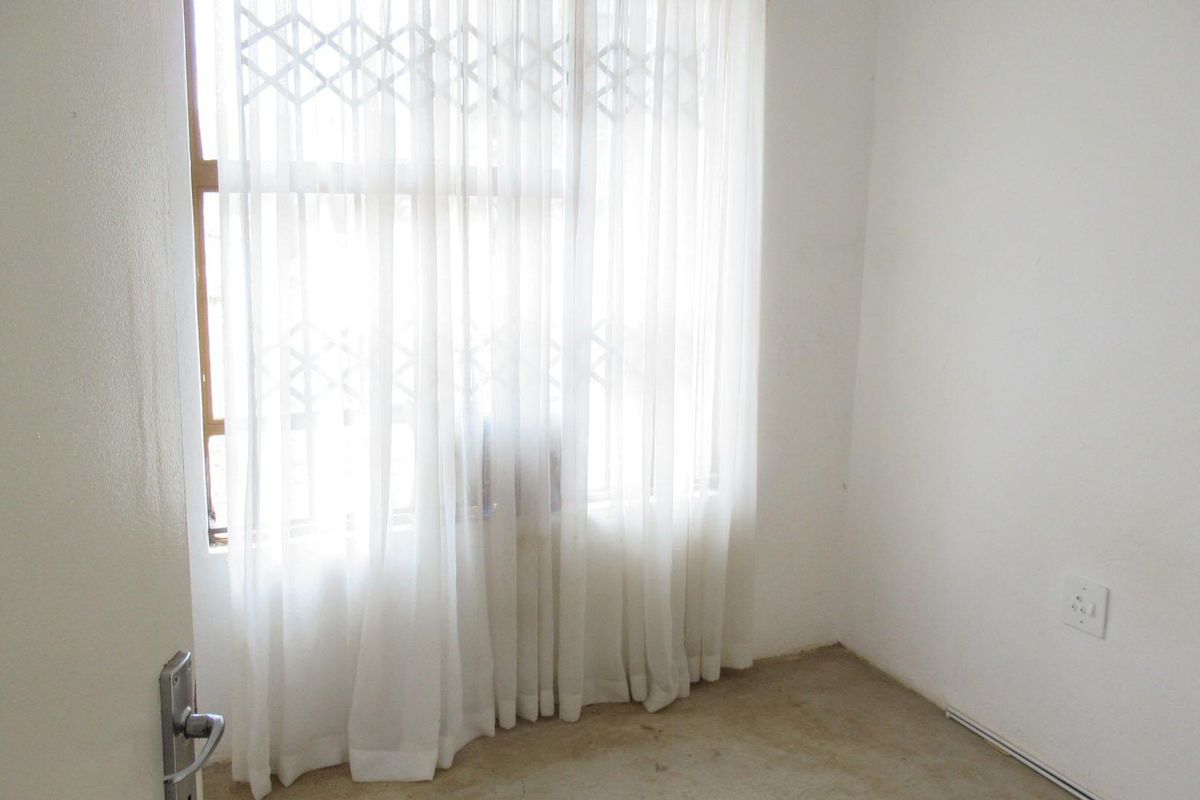 An empty bedroom featuring a window with a security gate and an electrical outlet, requiring flooring installation to complete the space.