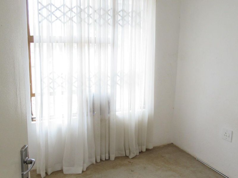 An empty bedroom featuring a window with a security gate and an electrical outlet, requiring flooring installation to complete the space.