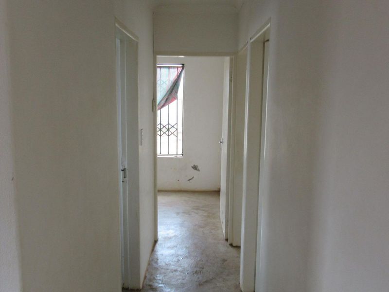 A vacant hallway with plain white walls and visible wear to the flooring and wall surfaces, leading towards a room with a security gate.