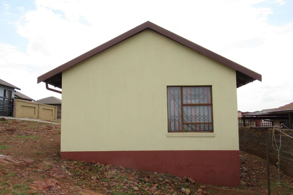 A side view of the house exterior featuring a single window with security bars, painted walls, and a plinth, set on an unlandscaped plot.