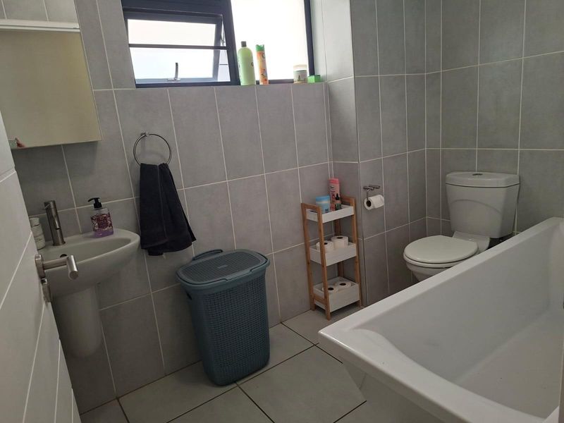 A functional bathroom featuring grey wall and floor tiles, a pedestal basin, a standard toilet, and a bathtub with a top-hung window.