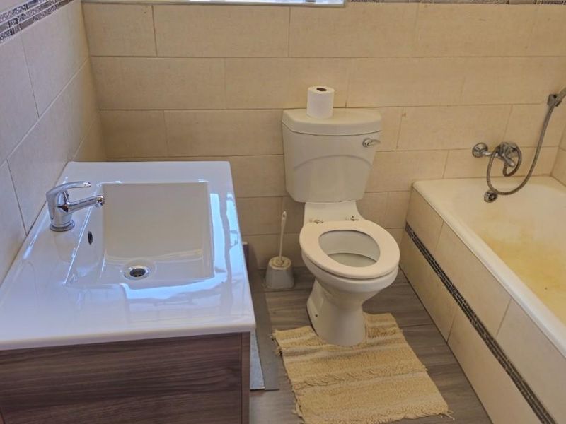 A compact bathroom featuring a modern vanity, a standard toilet, and a bathtub with shower mixer, finished with tiled walls and wood-look flooring.