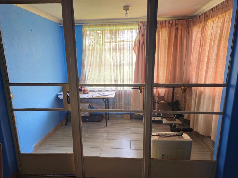 A small room viewed through glass panel doors, featuring tiled flooring and large windows with curtains, suitable for use as a home office or study.