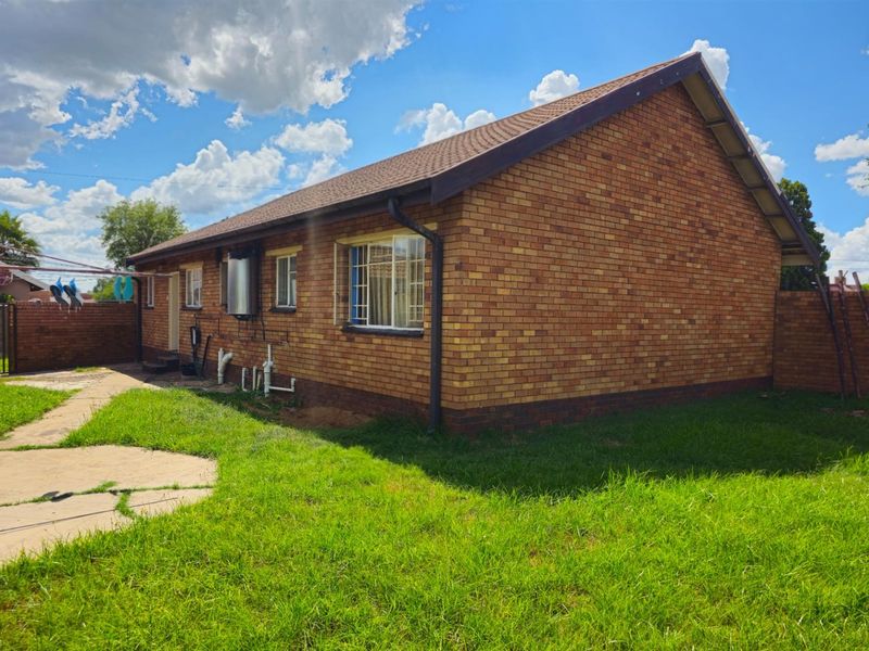 A view of the rear exterior of the brick house, featuring a tiled roof, concrete pathway, and surrounding lawn area.