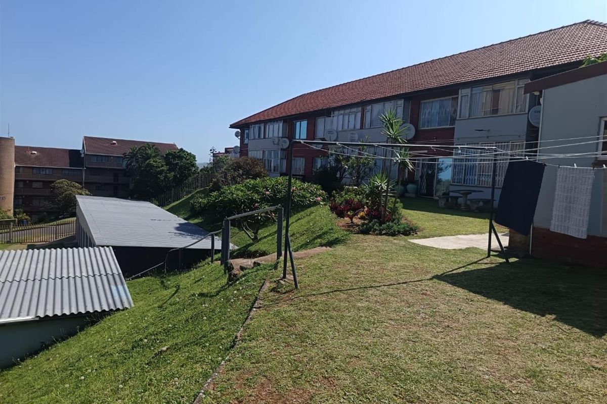 Rear view of the apartment complex showing the communal garden area, lawn, and adjacent building structures under a clear sky.