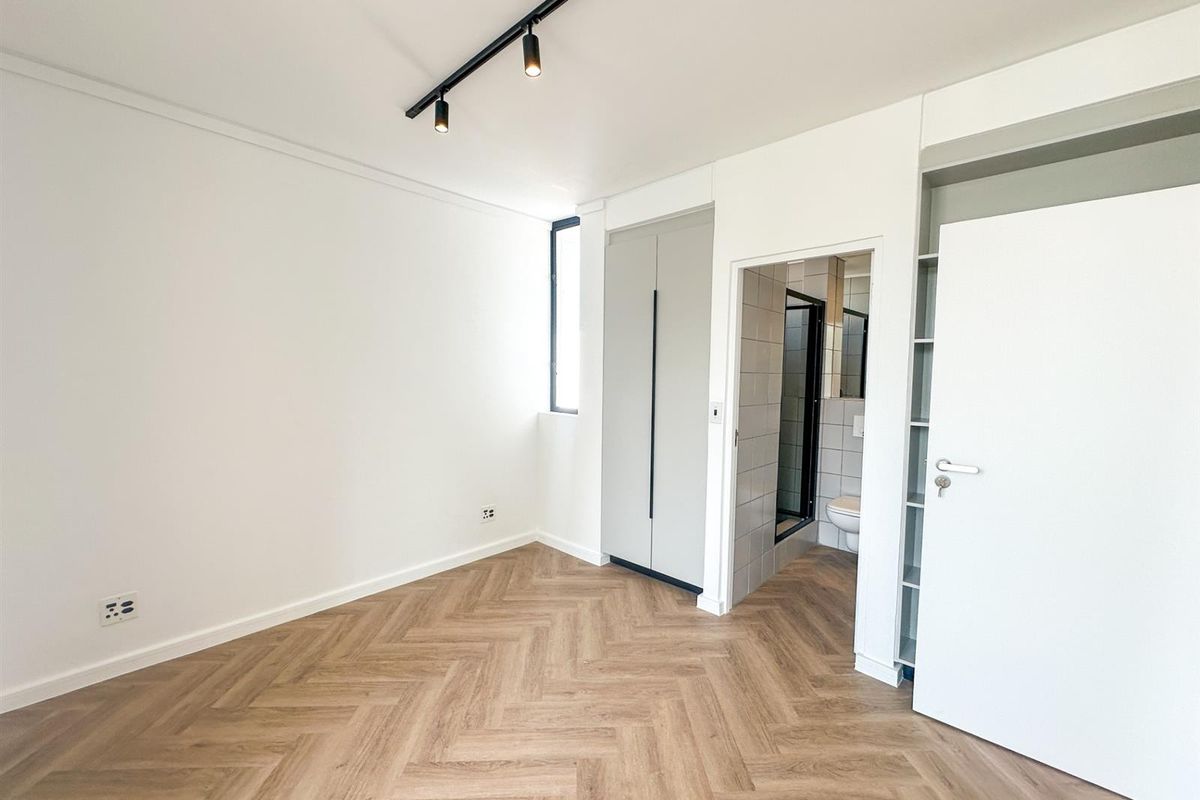 A bright, modern bedroom featuring herringbone wood-look flooring, built-in cabinetry, track lighting, and an en-suite bathroom.