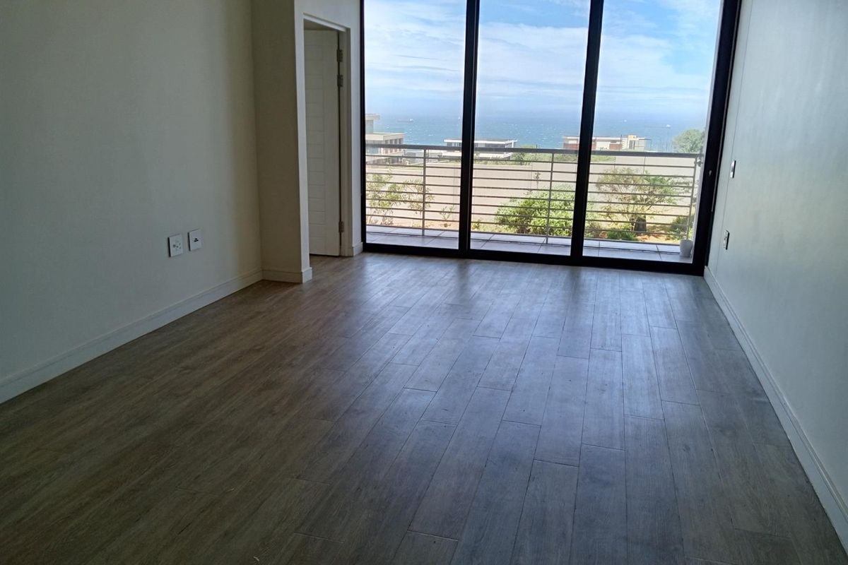 A bright, empty living room featuring modern wood-look tile flooring, large floor-to-ceiling windows, and direct access to a balcony with ocean views.