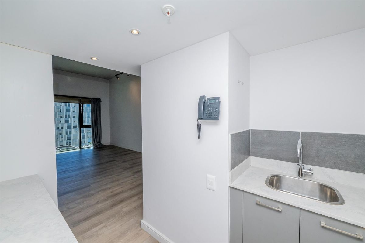 A compact kitchenette area featuring a stainless steel sink, grey cabinetry, and stone-look countertops, adjacent to an open-plan living space with wood-look flooring.