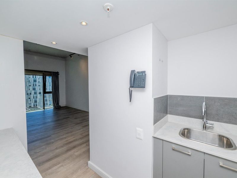 A compact kitchenette area featuring a stainless steel sink, grey cabinetry, and stone-look countertops, adjacent to an open-plan living space with wood-look flooring.