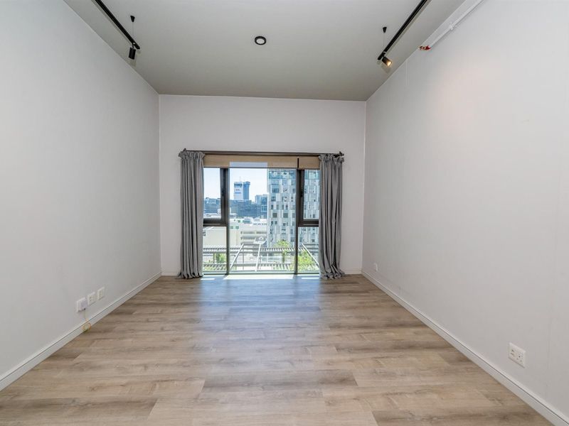 A bright, empty bedroom featuring modern light wood-look flooring, track lighting, and a large window offering an urban city view.