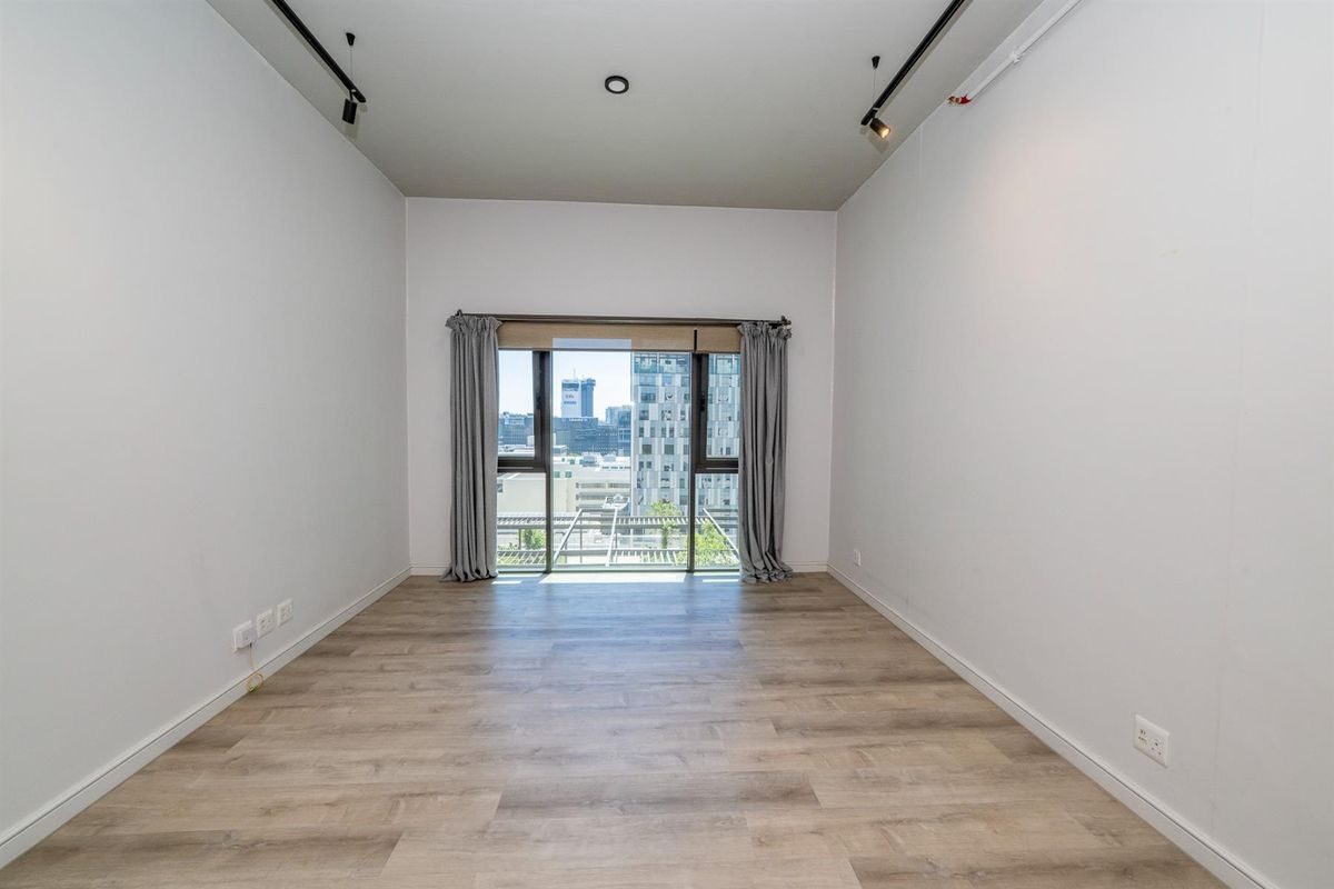A bright, empty bedroom featuring modern light wood-look flooring, track lighting, and a large window offering an urban city view.