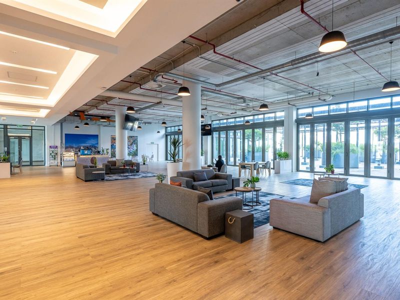Spacious open-plan communal living area featuring industrial-style exposed concrete ceilings, recessed lighting, wood-look flooring, and large glass sliding doors leading to an outdoor space.