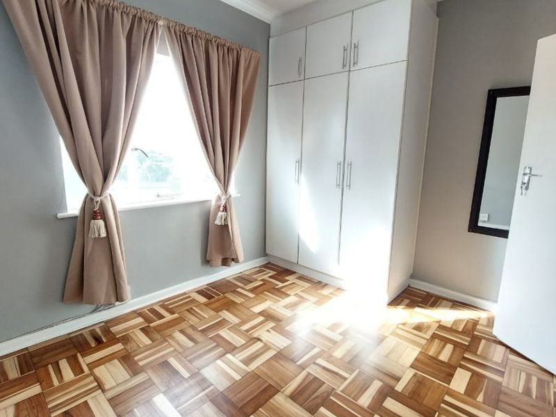 A bright, empty bedroom featuring well-maintained parquet flooring, built-in white cabinetry, and neutral grey walls, offering a clean and versatile living space.