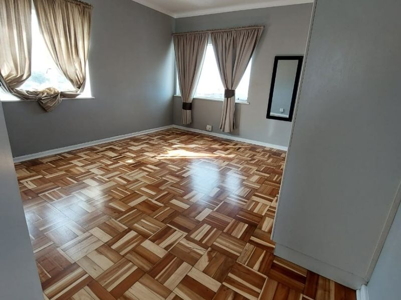 A spacious, empty bedroom featuring well-maintained parquet flooring, neutral grey walls, and natural light from two windows.