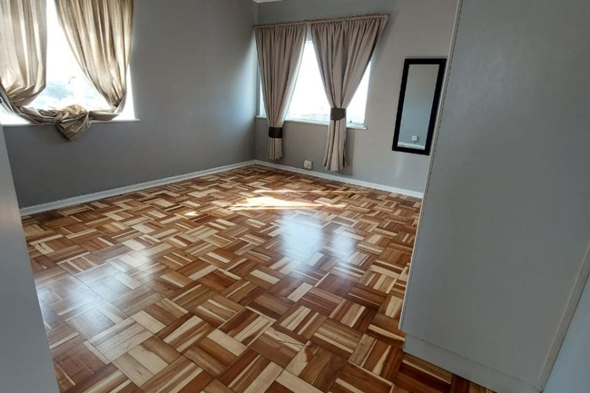 A spacious, empty bedroom featuring well-maintained parquet flooring, neutral grey walls, and natural light from two windows.