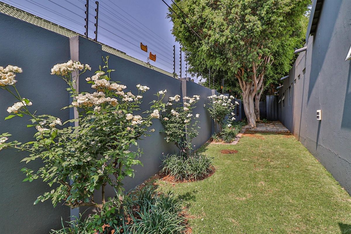 A well-maintained rear garden area featuring a high perimeter wall with integrated electric fencing, mature trees, and a paved pathway.