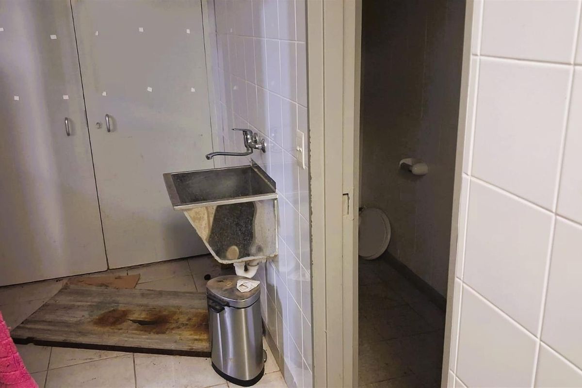 A utilitarian bathroom space featuring a wall-mounted stainless steel sink, tiled walls, and an adjacent toilet cubicle.