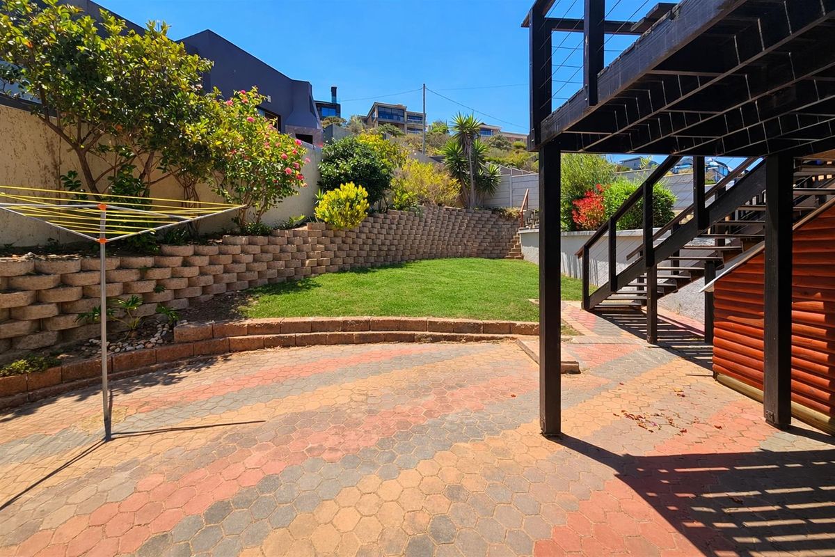 A rear garden featuring a paved patio area, tiered retaining walls, a lawn, and an elevated timber deck structure.