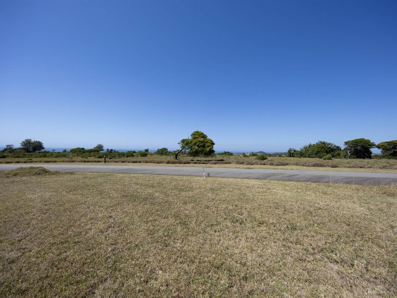 A view of a vacant residential plot featuring natural grass coverage and a paved road frontage with distant coastal views.