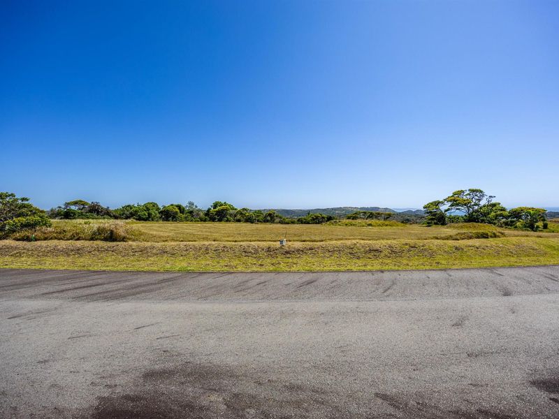A vacant residential plot of land featuring a gentle slope, natural grass cover, and distant landscape views, accessible from a paved road.