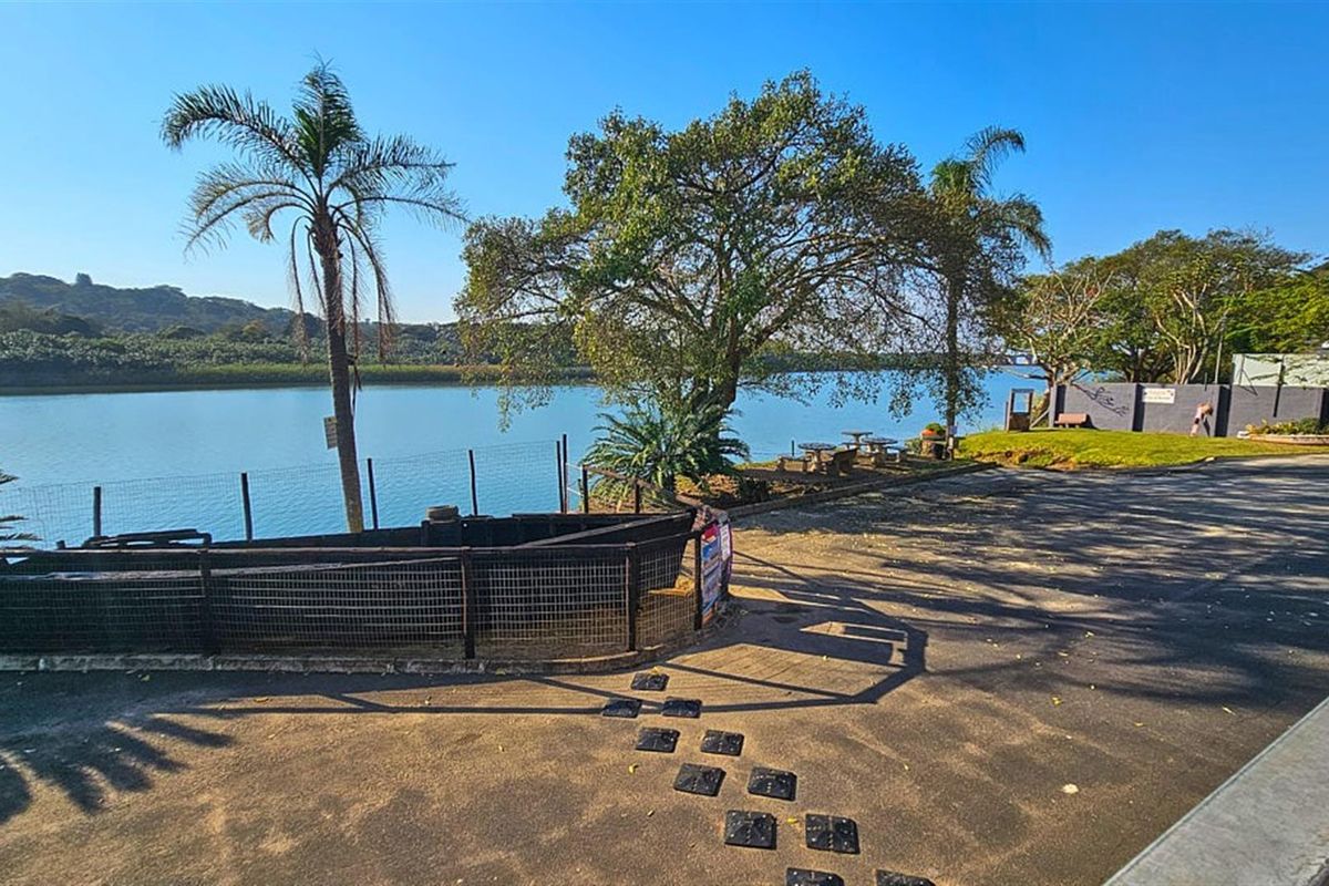 A scenic outdoor restaurant area featuring paved grounds, perimeter fencing, and mature trees overlooking a tranquil river view.