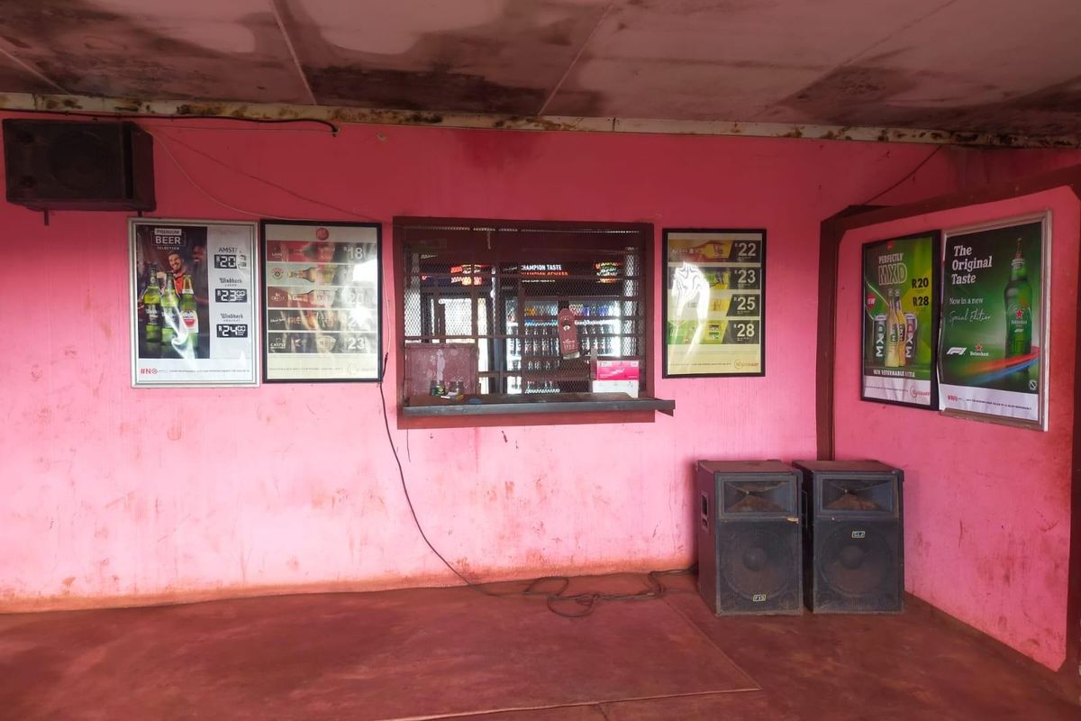 A commercial bar area featuring a serving hatch with security grille, pink painted walls, and a concrete floor, requiring maintenance and cosmetic updates.