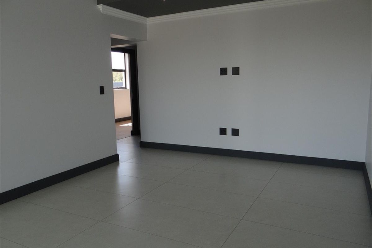 A bright, empty living room featuring modern large-format floor tiles, contrasting dark skirting boards, and recessed ceiling lighting.