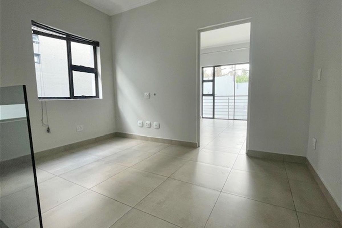 A bright, empty bedroom featuring neutral tiled flooring, modern aluminium-framed windows, and a doorway leading to an adjacent living space.