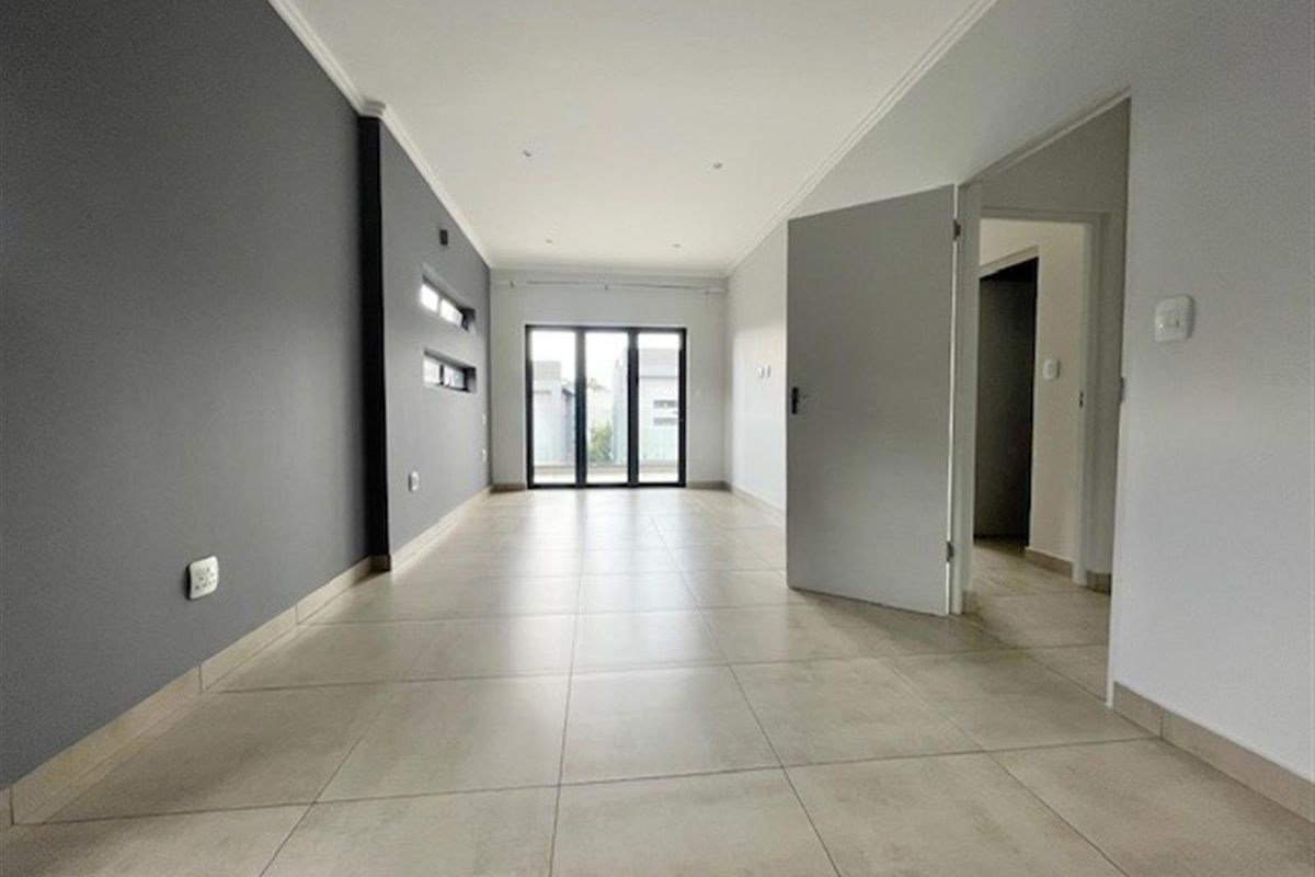 A bright, empty living room featuring large format floor tiles, a modern grey accent wall, recessed downlighting, and glass sliding doors leading to the exterior.