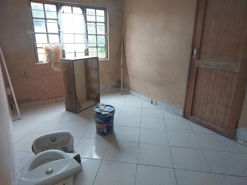 A vacant room undergoing renovation, featuring tiled flooring, a wooden door, and a window with security bars, currently in need of significant cosmetic repair.